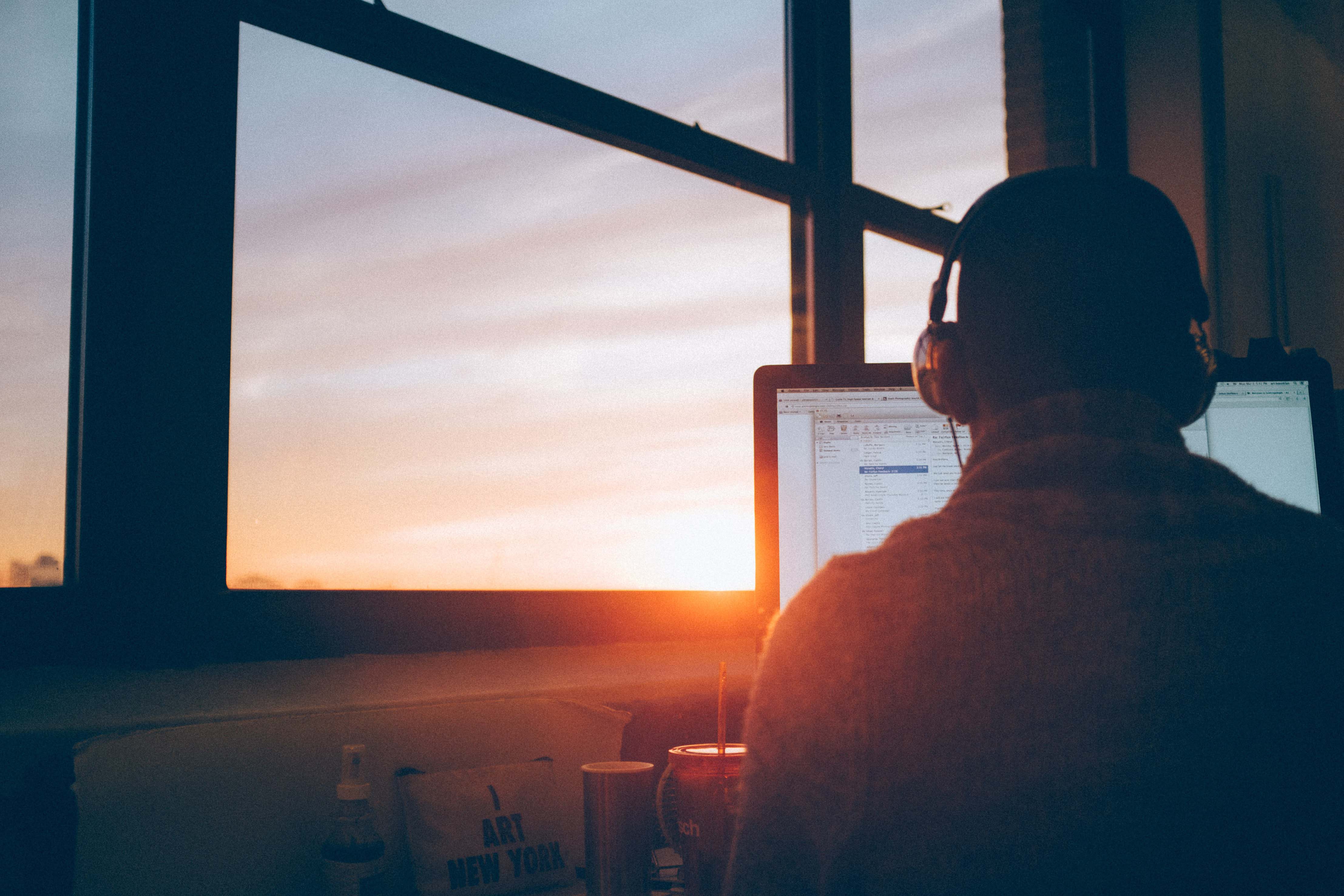 Man wearing headphones sitting in front of a computer with his back to the camera. He is sitting at a window and the sun is setting.