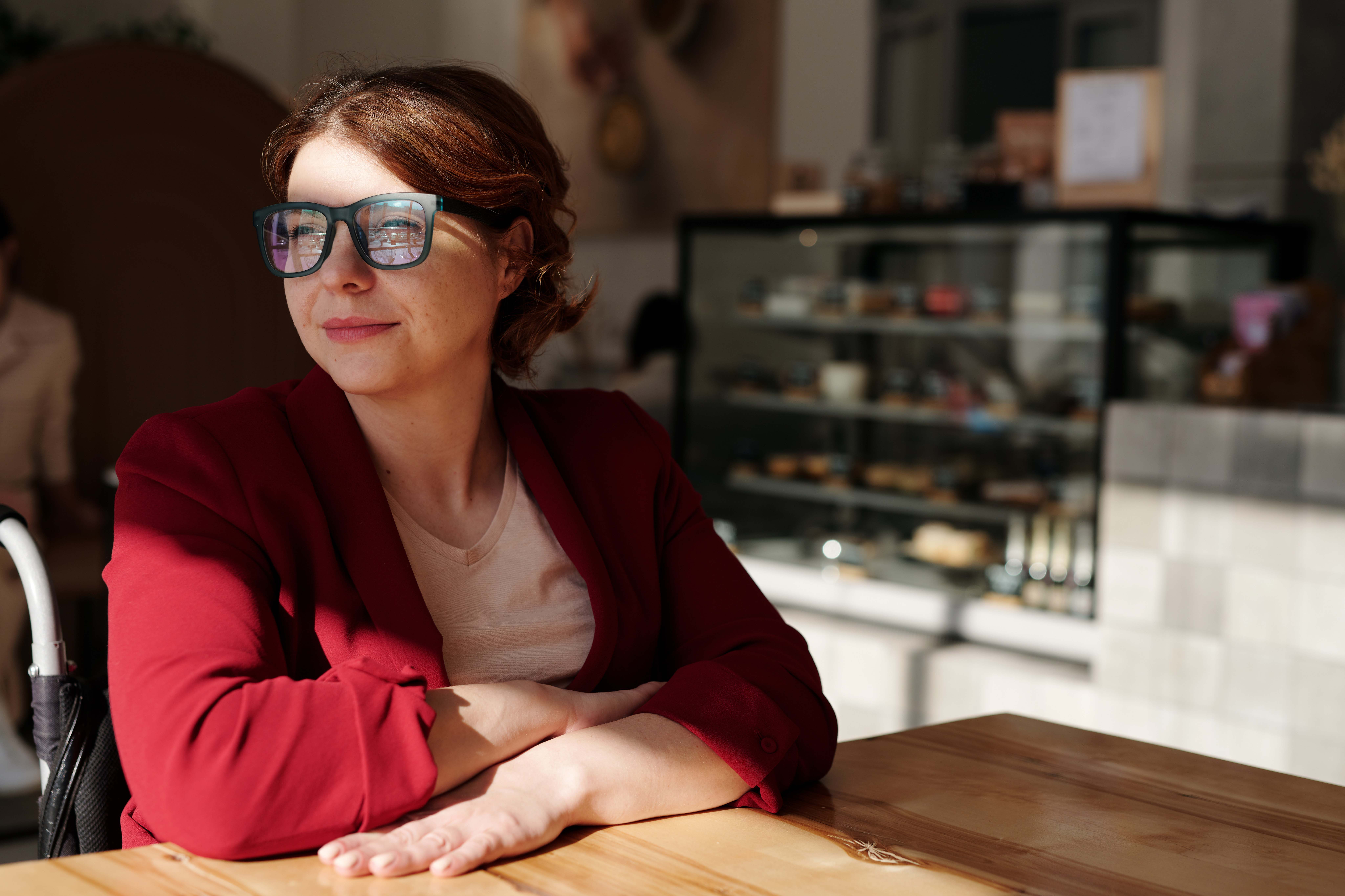 Woman in coffee shop looking towards a window with sun shining across her. She's wearing a red long sleeved jacket and black rimmed glasses.