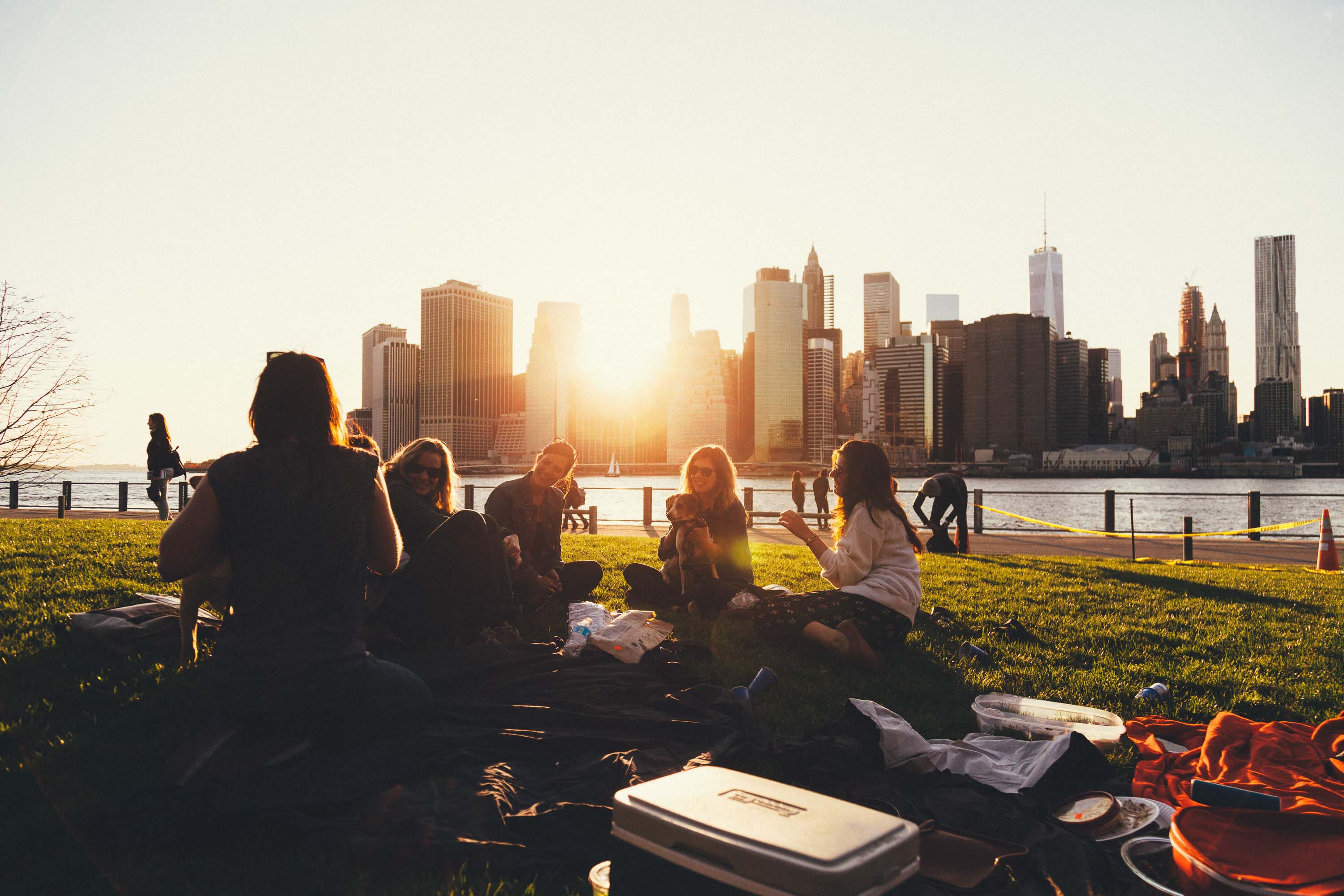 A group of 5 female friends are having a picnic on a grassy lawn. Two of the women have their dogs with them. In the background is a river and a city skyline, with the sun setting behind the buildings.