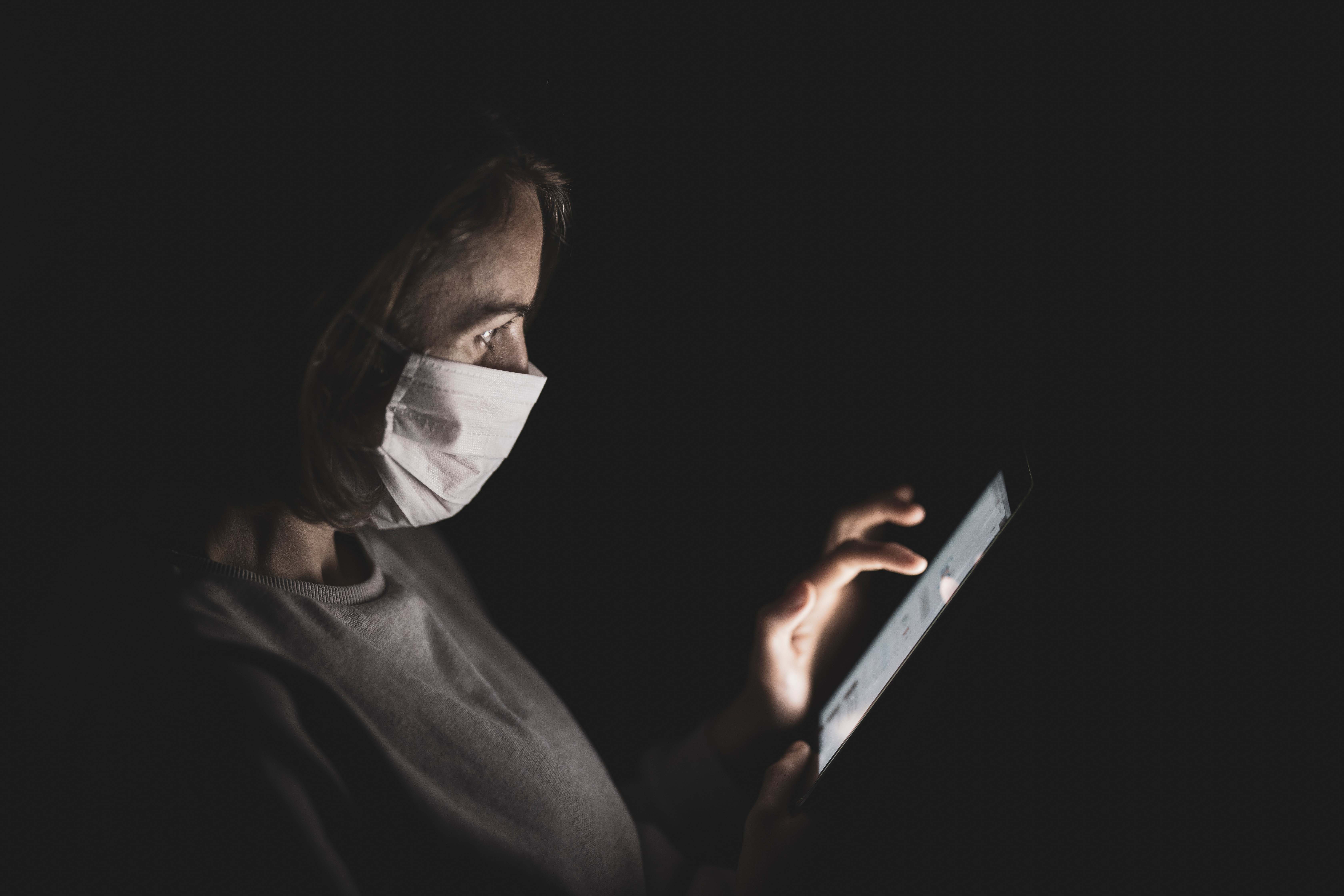Woman wearing a face mask for protection from coronavirus. She is sitting in the dark, illuminated from the light of a tablet that she is working on.